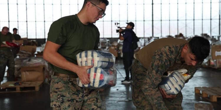 Two service members in camouflage uniforms carry packages of supplies in a large hangar.