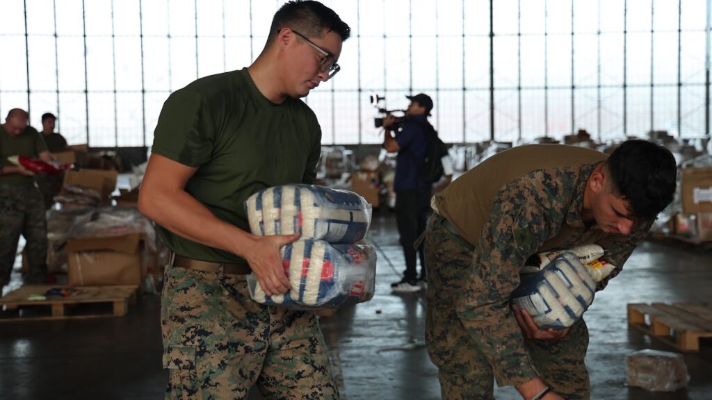 'When Times Get Tough, America Shows Up': US Ambassador to Panama Details Earthquake Recovery Efforts 1 Two service members in camouflage uniforms carry packages of supplies in a large hangar.